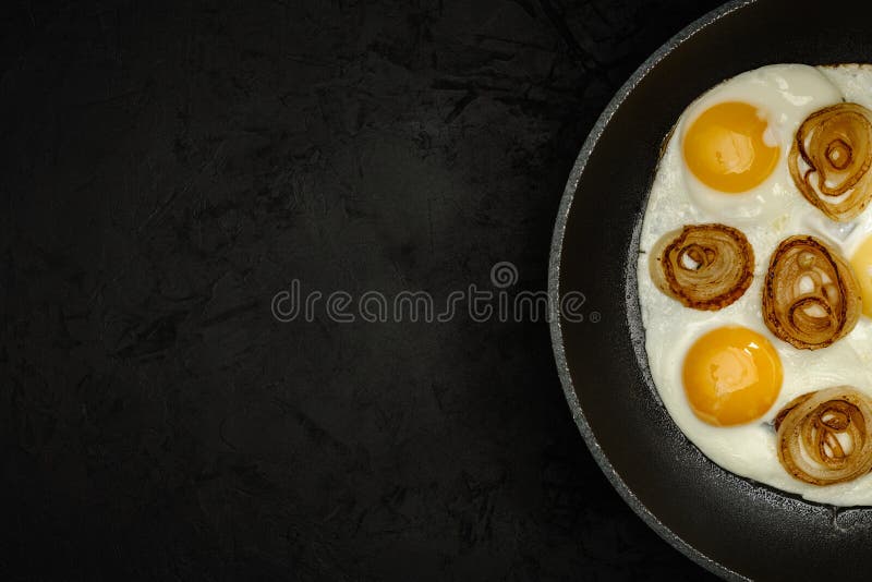 Frische Spiegeleier mit Zwiebelringen in einer Bratpfanne auf einem schwarzen strukturierten Gipshintergrund. Draufsicht. künstler stockbild