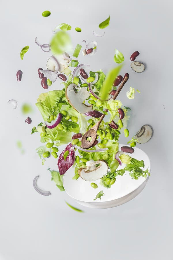 Frisk, grön sallad med svampar, njurbönor, ödem och lök som flyter över vit skål vid vit vägg.. flygmat arkivfoto