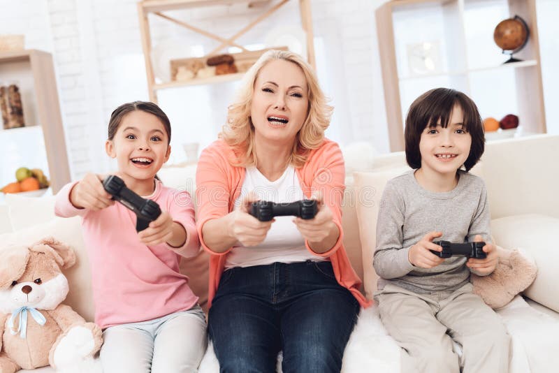 Frohe Großmutter mit den netten Enkelkindern, die auf der Spielkonsole sitzt auf Couch spielen lizenzfreie stockbilder Frohe Großmutter mit den netten Enkelkindern, die auf der Spielkonsole sitzt auf Couch spielen lizenzfreie stockbilder