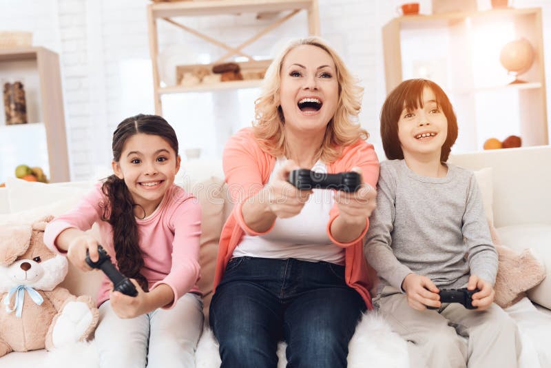 Frohe Großmutter mit den netten Enkelkindern, die auf der Spielkonsole sitzt auf Couch spielen lizenzfreie stockbilder Frohe Großmutter mit den netten Enkelkindern, die auf der Spielkonsole sitzt auf Couch spielen lizenzfreie stockbilder