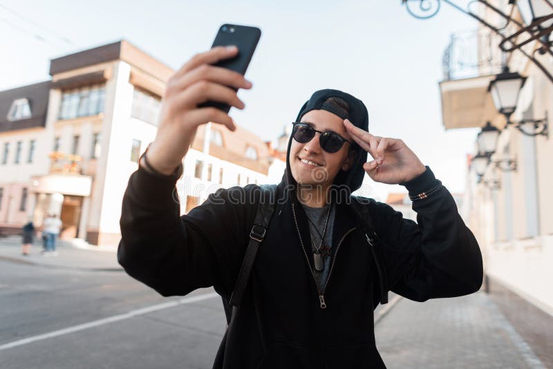Froher netter junger Hippie-Mann im modischen Hoodie, der in der dunklen Sonnenbrille in einer stilvollen Kappe mit einem Lächeln lizenzfreies stockfoto Froher netter junger Hippie-Mann im modischen Hoodie, der in der dunklen Sonnenbrille in einer stilvollen Kappe mit einem Lächeln lizenzfreies stockfoto