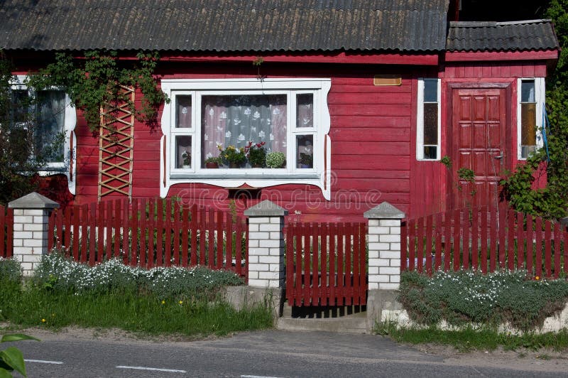 The front of red wooden house with white window, fence and gate. Beautiful flowers near the fence. The front of red wooden house with white window, fence and gate. Beautiful flowers near the fence.