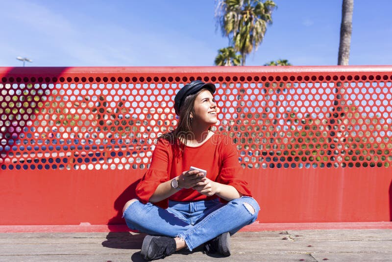Front view of a beautiful young woman wearing urban clothes sitting on a bridge floor while using a mobile phone outdoors in a stock images Front view of a beautiful young woman wearing urban clothes sitting on a bridge floor while using a mobile phone outdoors in a stock images