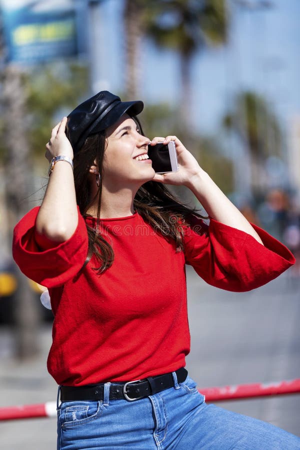 Front view of a beautiful young woman wearing urban clothes sitting on a metallic fence while using a mobile phone outdoors in the stock photography Front view of a beautiful young woman wearing urban clothes sitting on a metallic fence while using a mobile phone outdoors in the stock photography