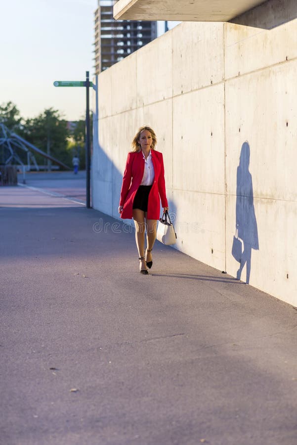 Front view of a elegant woman wearing red jacket, skirt and holding a white handbag while walking in the street in a sunny day