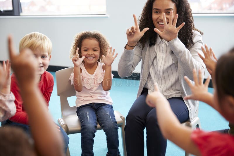 Front view of infant school children sitting on chairs in a circle in the classroom, holding up their hands and learning to count.