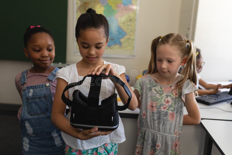 Front view of schoolgirl with curious classmates holding a virtual reality headset in classroom of elementary school. Front view of schoolgirl with curious classmates holding a virtual reality headset in classroom of elementary school