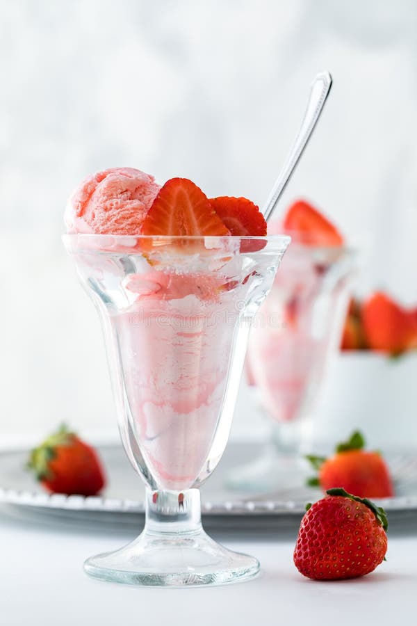 A front view of a strawberry ice cream sundae with another one on a metal tray in behind and a bowl of strawberries.