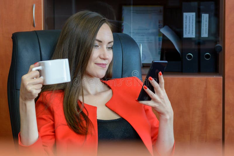 Frontowego widoku portret młoda kobieta w czerwonym garnituru obsiadaniu w rzemiennego krzesła pić kawowy i używać smartphone zdjęcia stock Frontowego widoku portret młoda kobieta w czerwonym garnituru obsiadaniu w rzemiennego krzesła pić kawowy i używać smartphone zdjęcia stock