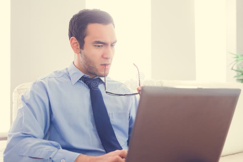 Frowning man biting his eyeglasses and using a laptop