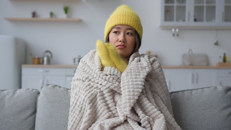 Frozen Asian chinese woman in warm hat and mittens rubbing freezing hands sit in cold kitchen frosty temperature heating