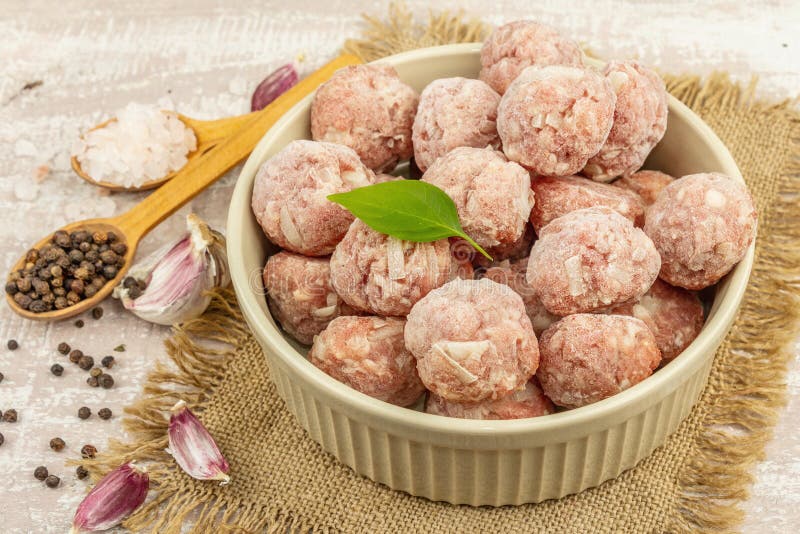 Frozen meatballs with spices, basil leaves, and garlic cloves in a ceramic bowl