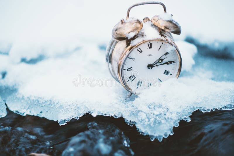 Frozen time. A clock on an ice next to the water. Extreme weather situation. Snow falling on a clock in a nature.