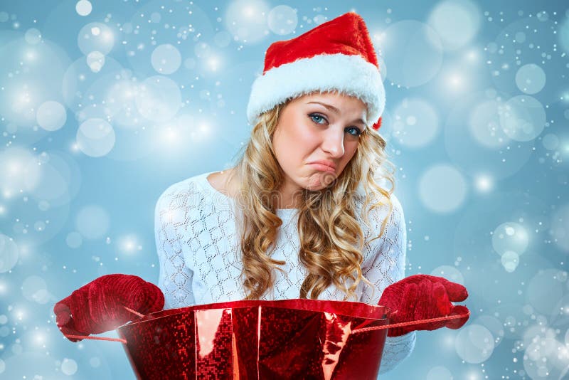 Frustrated and annoyed beautiful young woman in Santa Claus hat with a gift on a blue background