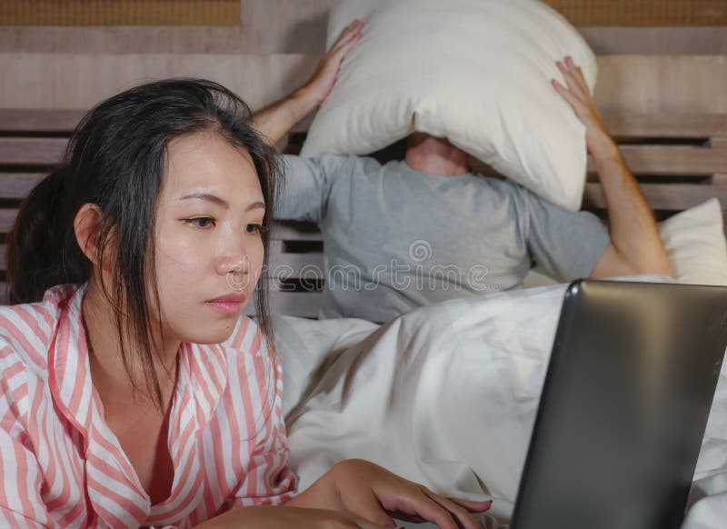 Frustrated Husband Moody in Bed Ignored by His Workaholic Asian Wife or Internet Social Media Addict Girlfriend Using Laptop in Stock Photo