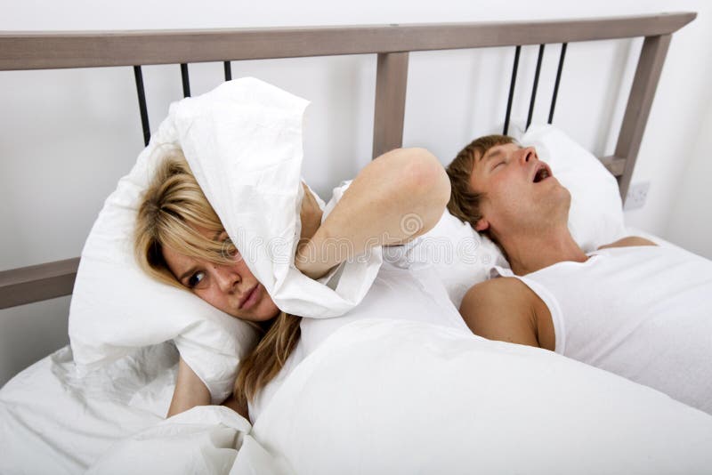 Frustrated woman covering ears with pillow while man snoring in bed