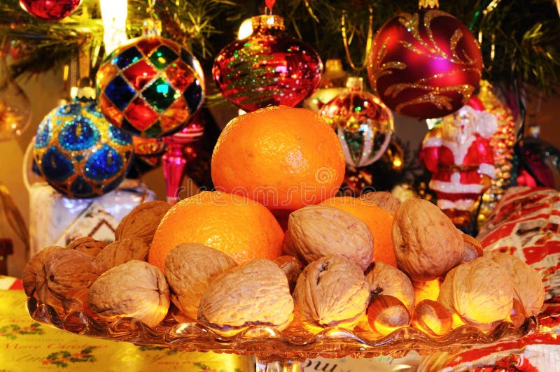 Glass plate stand topped with walnuts, hazelnuts and mandarin oranges with Christmas presents under tree to the rear, England, UK, Western Europe. Glass plate stand topped with walnuts, hazelnuts and mandarin oranges with Christmas presents under tree to the rear, England, UK, Western Europe.