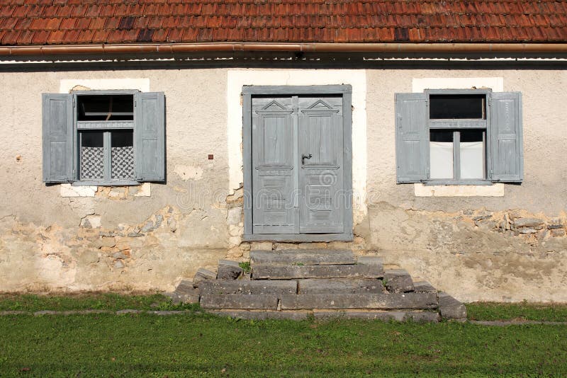 Främre ingång till det gamla övergav huset med gräs- och stenmoment framme av trädörrar som omges med två fönster med öppet arkivfoton Främre ingång till det gamla övergav huset med gräs- och stenmoment framme av trädörrar som omges med två fönster med öppet arkivfoton