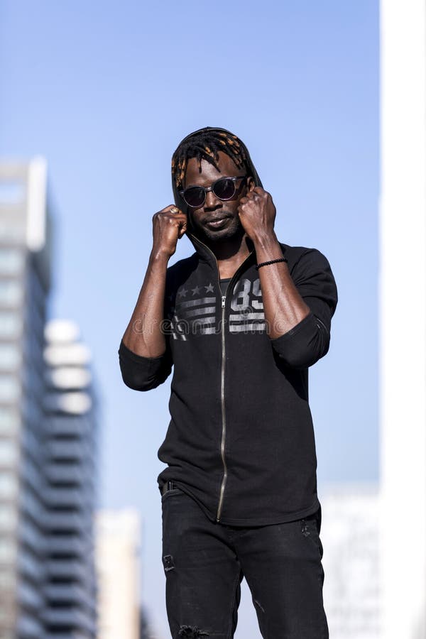 Främre sikt av en ung svart afrikansk man som bär tillfällig kläder och solglasögon som står i gatan, medan se kameran i a royaltyfri fotografi Främre sikt av en ung svart afrikansk man som bär tillfällig kläder och solglasögon som står i gatan, medan se kameran i a royaltyfri fotografi