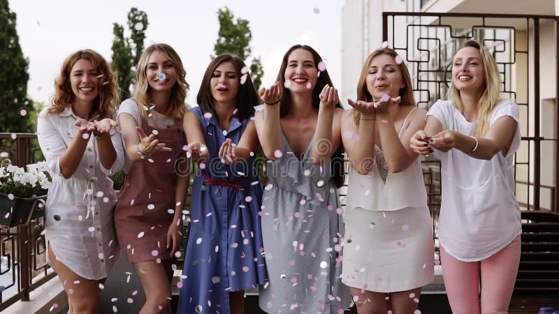 Främre sikt av sex attraktiva caucasian unga kvinnor som playfully tillsammans blåser ljusa kulöra konfettier från deras händer