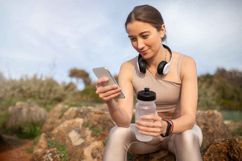Fröhliche Jahrtausendvikasische Frauensportlerin trägt drahtlose Kopfhörer, die telefonisch chatten, auf Stein stockfotografie