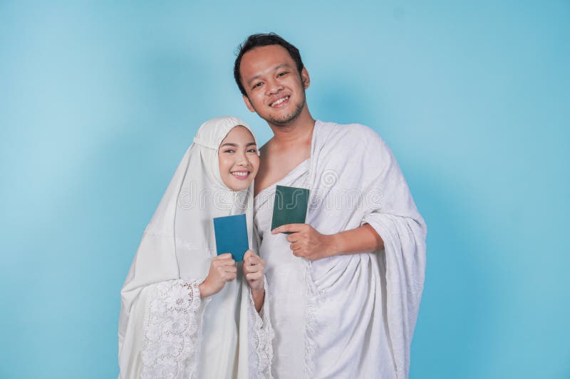 Fröhliches asiatisches Muslimpaar in ihram und Gebetskleid, das mit einem Pass, abgeschirmt durch blauen Hintergrund posiert. Hads stockbild