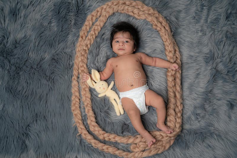 Full body picture of the boy lying on a grey fur having a rabbit toy, looking curious at the viewer