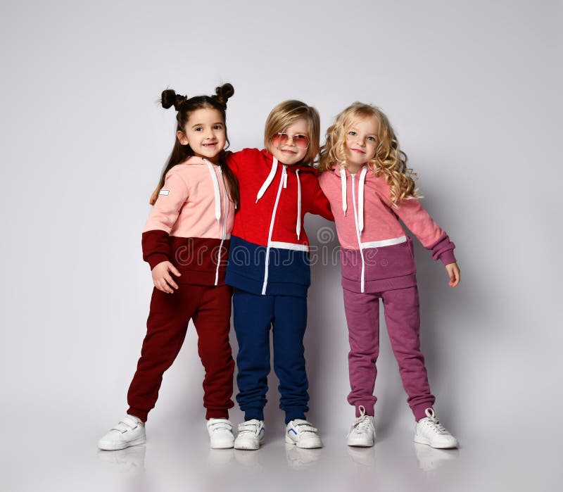 Full growth portrait of three happy smiling kids best friends team blonde and brunette girls and blond boy in modern two-colored hoodies and pants stands together hugging. Full growth portrait of three happy smiling kids best friends team blonde and brunette girls and blond boy in modern two-colored hoodies and pants stands together hugging