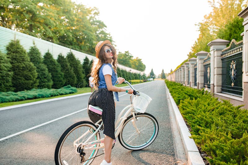 Full-length photo of cute girl with long curly hair in sunglasses driving a bike on road. She wears long skirt, jerkin