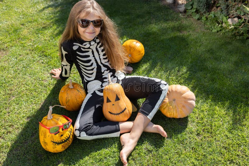 girl in a black skeleton costume and dark glasses sitting on the grass with orange pumpkins around her