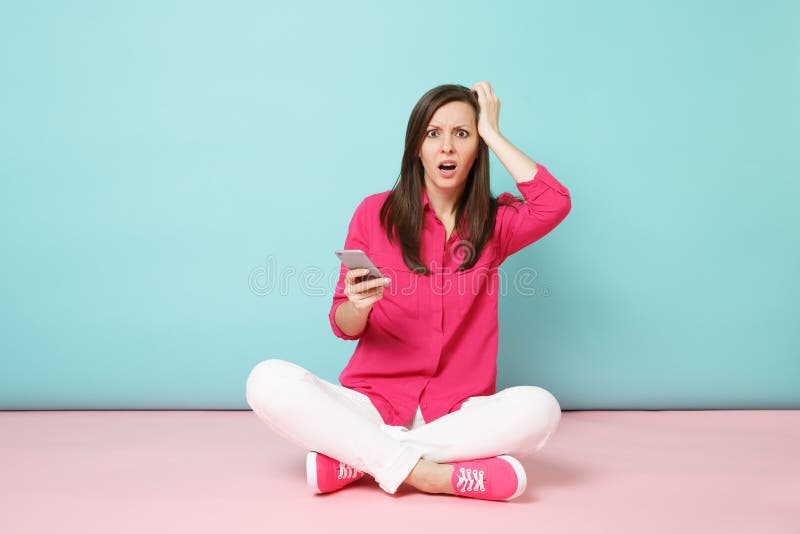 Full length portrait of young fun woman in rose shirt, white pants sit on floor hold cellphone isolated on bright pink