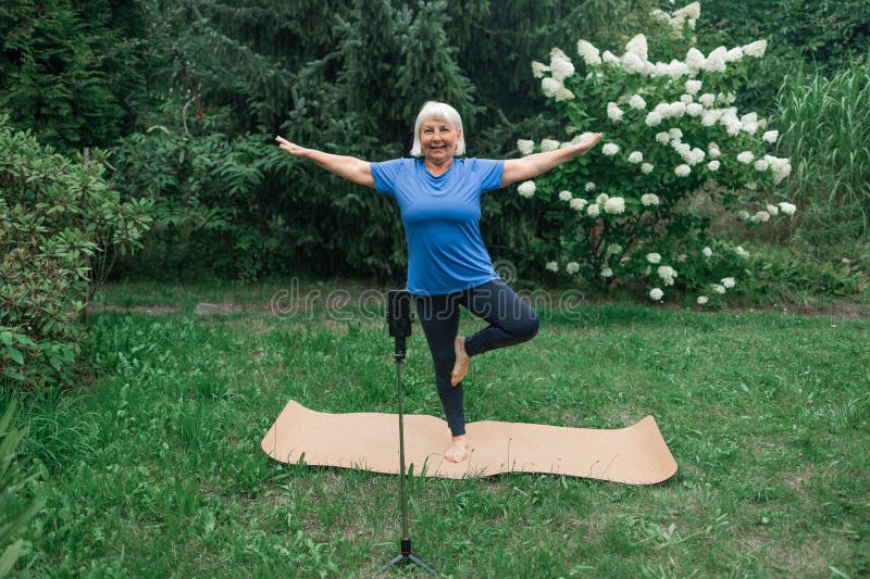 Yoga at park.Full length shot of happy energetic mature 50s woman in casual clothes exercising outdoors, practicing yoga on mat, standing in warrior ii pose. Age, wellness and health. Healthy lifestyle