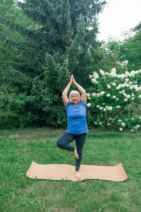 Yoga at park.Full length shot of happy energetic mature 50s woman in casual clothes exercising outdoors, practicing yoga on mat, standing in warrior ii pose. Age, wellness and health. Healthy lifestyle