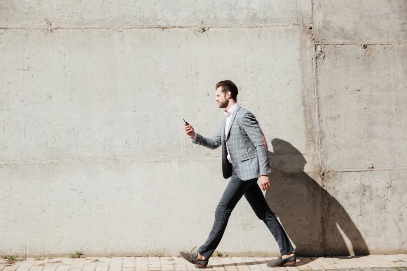 Full length side view portrait of a young man in jacket walking and using mobile phone infront of a wall outdoors. Full length side view portrait of a young man in jacket walking and using mobile phone infront of a wall outdoors