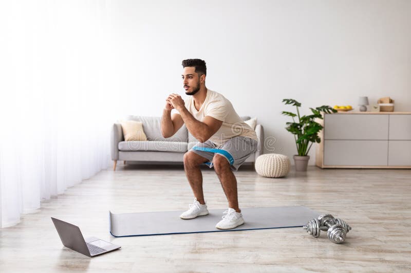 Full length of young Arab guy working out in front of laptop computer at home, following online video tutorial indoors. Millennial middle Eastern guy training with personal coach via internet. Laptop personal computer stock images, royalty-free photos and pictures