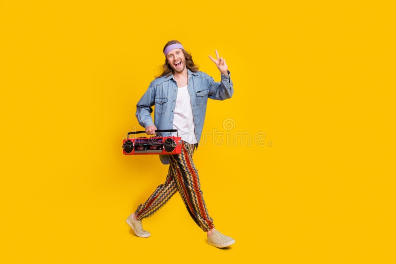 Full size profile photo of cool young man boombox show v-sign walk empty space wear denim shirt isolated on yellow color background. Full size profile photo of cool young man boombox show v-sign walk empty space wear denim shirt isolated on yellow color background.