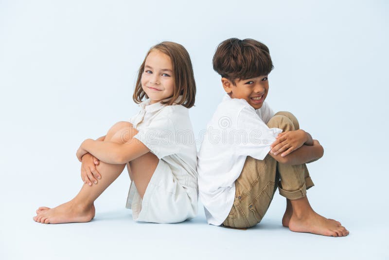 Fun portrait of awkwardly smiling boy and girl sitting back to back on the floor. against bluish white background. Both barefoot.