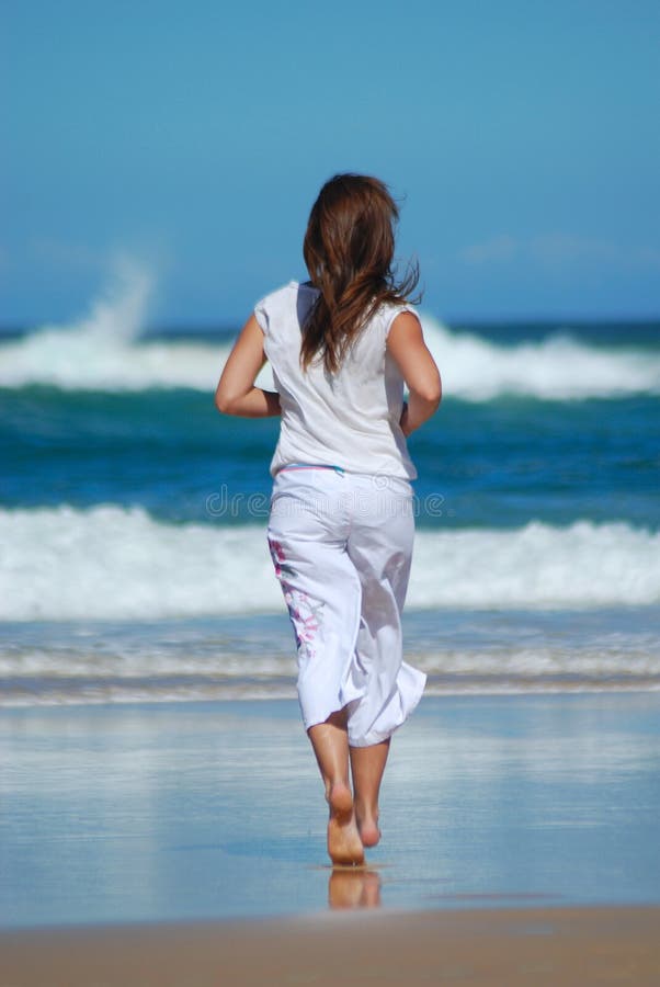 Back view of an active caucasian white fitness woman in white clothes running in the sand of the beach towards the blue sea. Back view of an active caucasian white fitness woman in white clothes running in the sand of the beach towards the blue sea