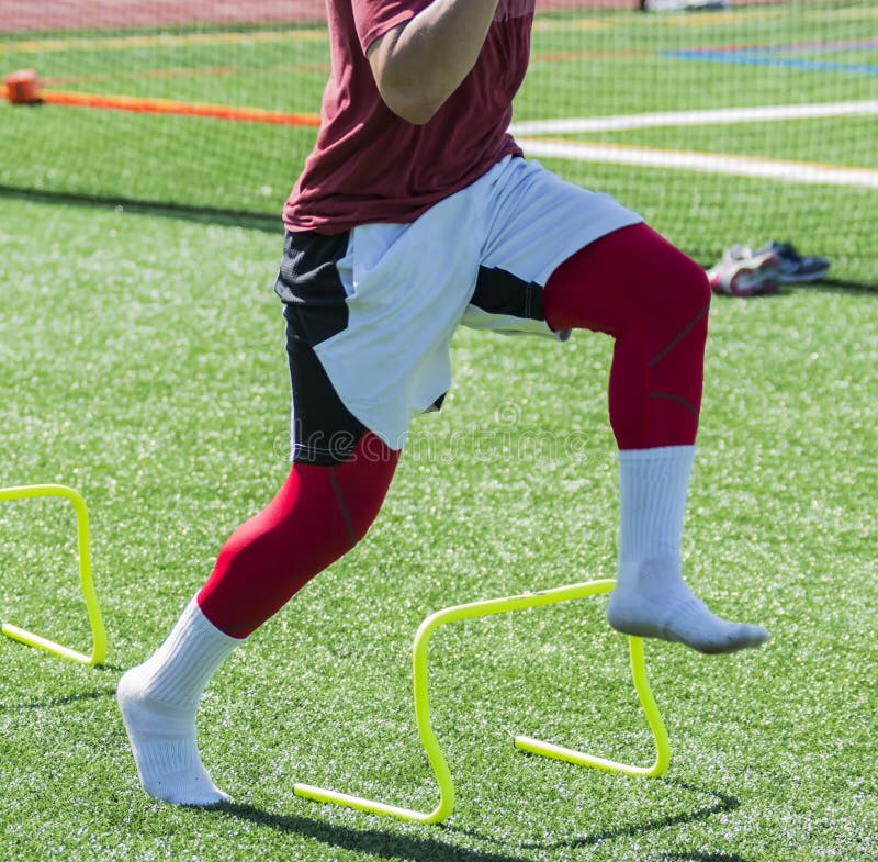 An athlete performs speed and agility drills by running over yellow mini hurdles wearing socks on a green turf field. An athlete performs speed and agility drills by running over yellow mini hurdles wearing socks on a green turf field.