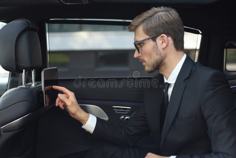 Fundersam ung affärsman som sitter i den luxe bilen och använder hans minnestavla arkivfoton Fundersam ung affärsman som sitter i den luxe bilen och använder hans minnestavla arkivfoton