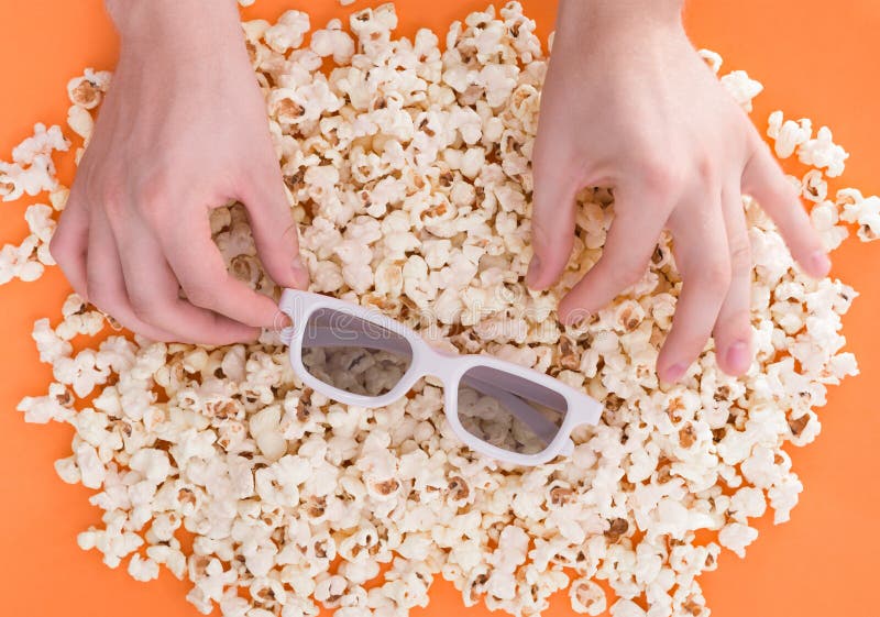 Background. Men`s hands on the background of a popcorn, one holds 3d glasses, the second takes popcorn, isolated on a blackened background, a view from above. Flat lay. Copyspace. Cinema Concept. Background. Men`s hands on the background of a popcorn, one holds 3d glasses, the second takes popcorn, isolated on a blackened background, a view from above. Flat lay. Copyspace. Cinema Concept