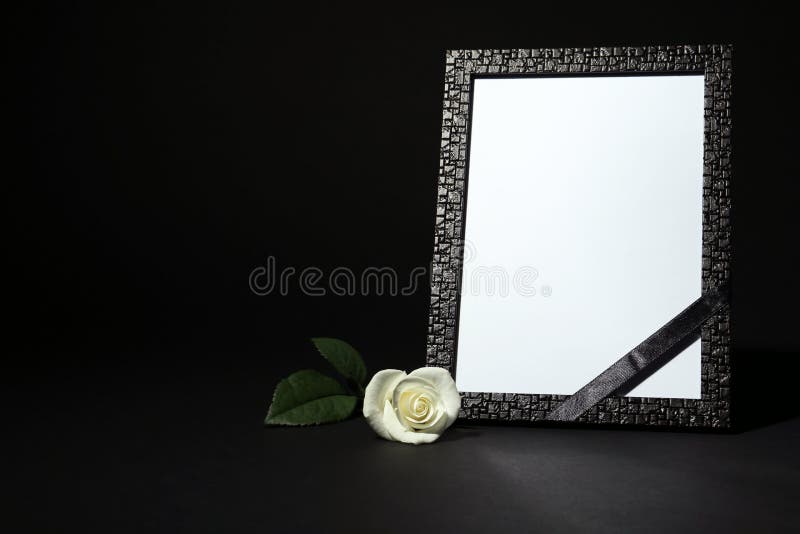 Funeral photo frame with ribbon and white rose on table against black background. Space for design