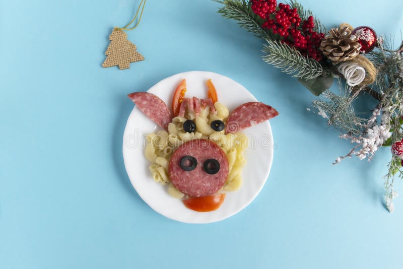 Funny Breakfast for children in the form of bull`s face made of pasta, sausage, bacon, olives and fresh tomatoes on a blue background. Plate with creative food art Breakfast for children with a Christmas tree branch and a cone Top view