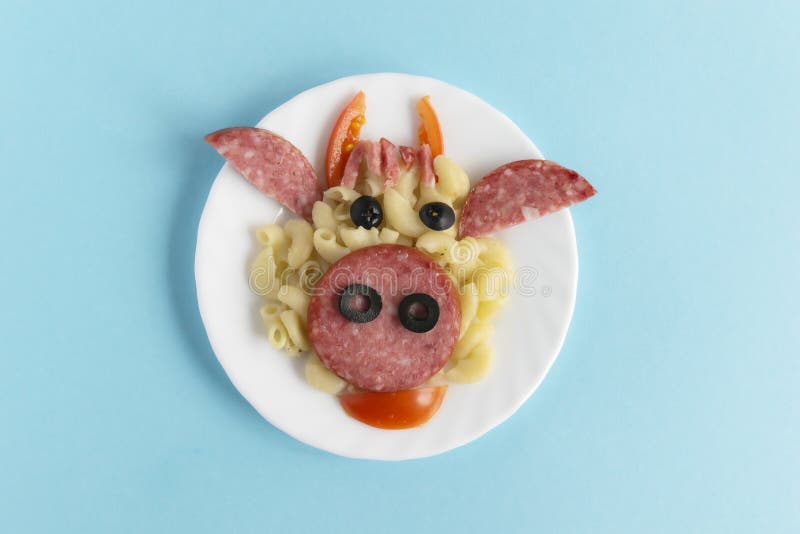 Funny Breakfast, lunch or dinner for children in the form of a bull`s face made of pasta, sausage, bacon, olives and fresh tomatoes on a blue background. Instruction how to make creative food art breakfast for kids. Step 8. Top view. Funny Breakfast, lunch or dinner for children in the form of a bull`s face made of pasta, sausage, bacon, olives and fresh tomatoes on a blue background. Instruction how to make creative food art breakfast for kids. Step 8. Top view