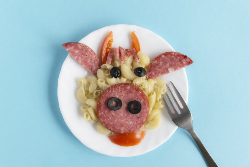 Funny Breakfast, lunch or dinner for children in the form of a bull`s face made of pasta, sausage, bacon, olives and fresh tomatoes on a blue background. Plate with creative food art Breakfast for children with a fork. Top view. Funny Breakfast, lunch or dinner for children in the form of a bull`s face made of pasta, sausage, bacon, olives and fresh tomatoes on a blue background. Plate with creative food art Breakfast for children with a fork. Top view