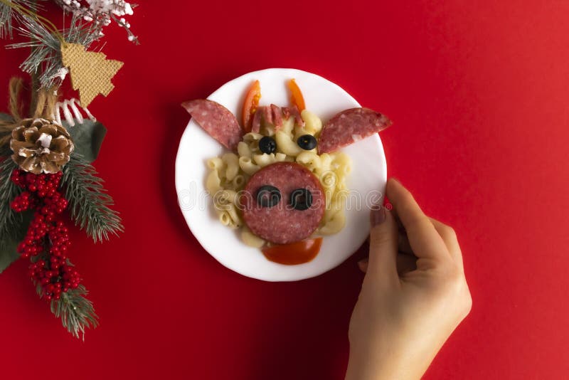 Funny Breakfast for children in the form of bull`s face made of pasta, sausage, bacon, olives and fresh tomatoes on a red background. Female hand hold plate with creative food art Breakfast for children with a Christmas tree branch and a cone Top view. Funny Breakfast for children in the form of bull`s face made of pasta, sausage, bacon, olives and fresh tomatoes on a red background. Female hand hold plate with creative food art Breakfast for children with a Christmas tree branch and a cone Top view