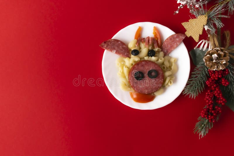 Funny Breakfast for children in the form of bull`s face made of pasta, sausage, bacon, olives and fresh tomatoes on a red background. Plate with creative food art Breakfast for children with a Christmas tree branch and a cone Top view