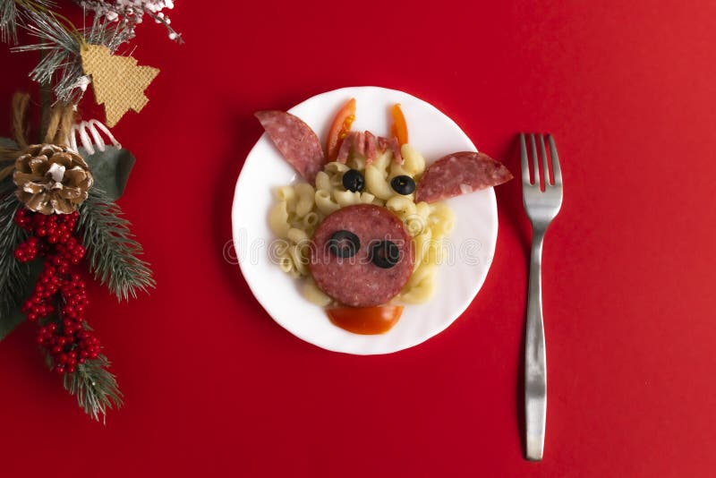 Funny Breakfast for children in the form of bull`s face made of pasta, sausage, bacon, olives and fresh tomatoes on a red background. Plate with creative food art Breakfast for children with with a fork and Christmas tree branch and a cone Top view