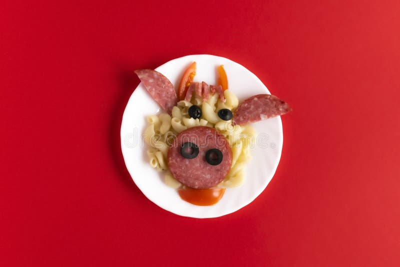 Funny Breakfast, lunch or dinner for children in the form of a bull`s face made of pasta, sausage, bacon, olives and fresh tomatoes on a red background. Instruction how to make creative food art breakfast for kids. Top view. Funny Breakfast, lunch or dinner for children in the form of a bull`s face made of pasta, sausage, bacon, olives and fresh tomatoes on a red background. Instruction how to make creative food art breakfast for kids. Top view