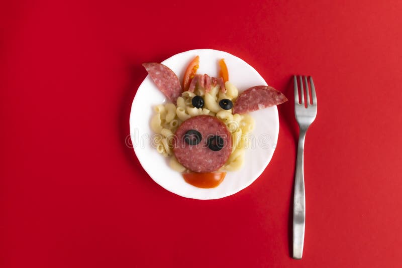 Funny Breakfast, lunch or dinner for children in the form of a bull`s face made of pasta, sausage, bacon, olives and fresh tomatoes on a red background. Plate with creative food art Breakfast for children with a fork. Top view. Funny Breakfast, lunch or dinner for children in the form of a bull`s face made of pasta, sausage, bacon, olives and fresh tomatoes on a red background. Plate with creative food art Breakfast for children with a fork. Top view